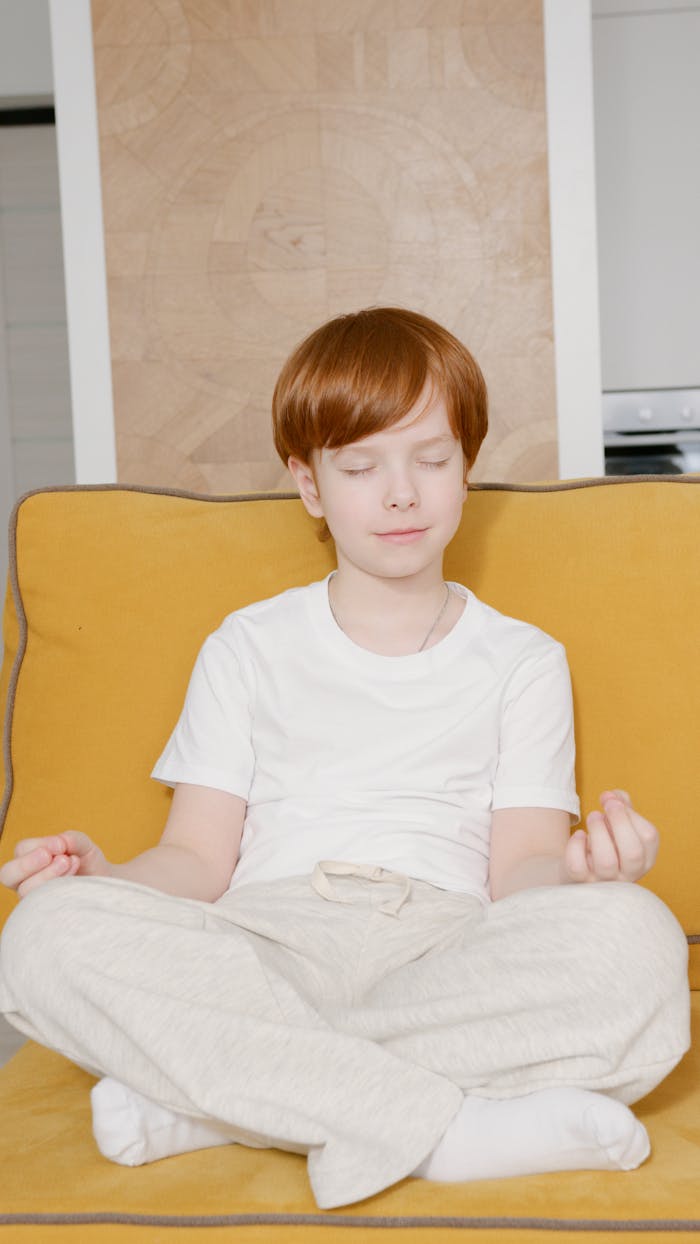 A red-haired child sits in a lotus pose meditating on a yellow sofa indoors.